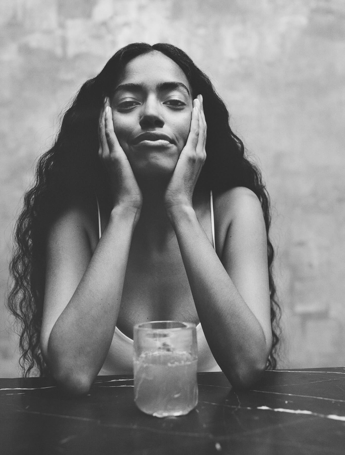 Black and white photo of a woman with hands on her face, sitting at a table with a glass.