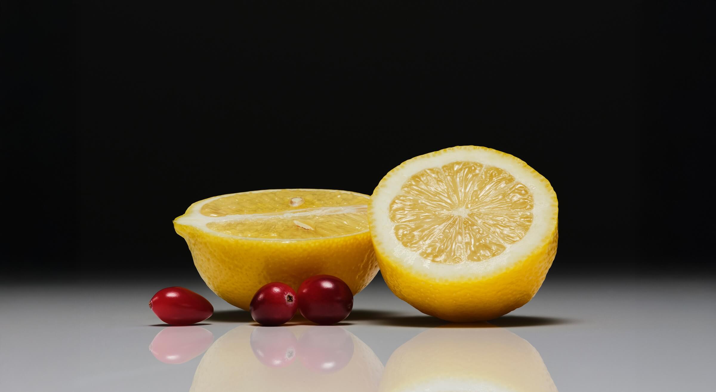 Two lemons, one whole and one halved, with two berries on a reflective surface against a black background.