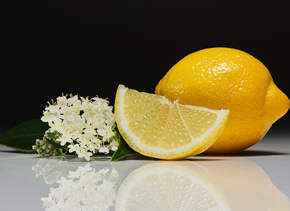 Two whole lemons and a lemon wedge with elderflower on a reflective surface against a black background