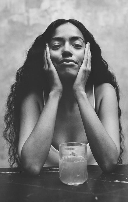 Black and white photo of a woman with hands on her face, sitting at a table with a glass of water.