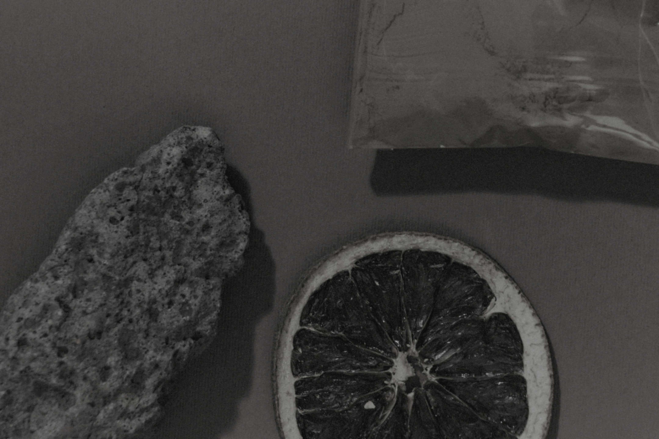 Black and white photo showing a dried citrus slice, a rough stone, and a plastic bag on a flat surface.