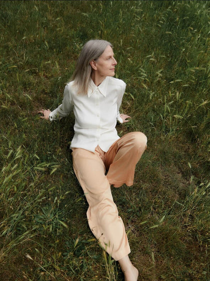Elegant mature woman in white blouse and peach pants sitting in a grassy field.